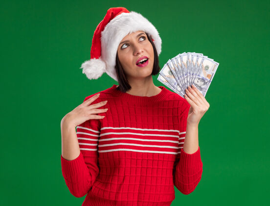 帽子印象深刻的年轻女孩戴着圣诞帽拿着钱抚摸着肩膀看着绿色的墙上孤立圣诞节穿着圣诞老人