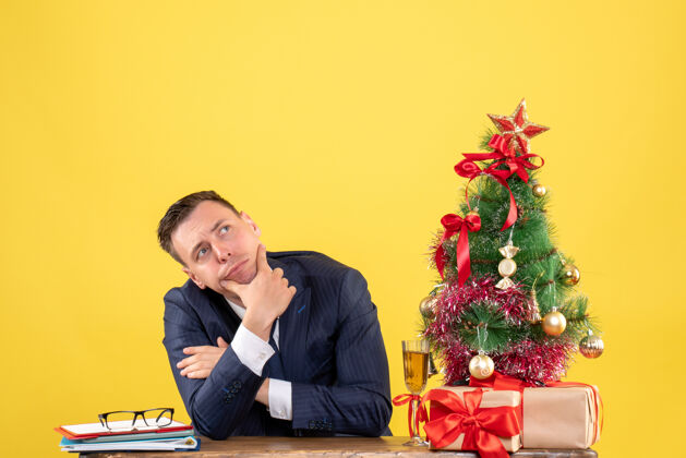 体贴的男人前视图：体贴的男人坐在圣诞树旁的桌子上 黄色的礼物商人坐着西装