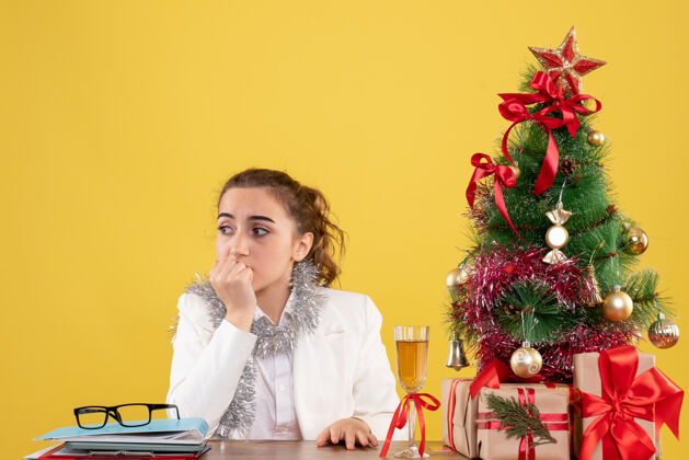 坐着正面图：女医生坐在黄色背景的桌子后面 还有圣诞树和礼品盒成人情感女医生