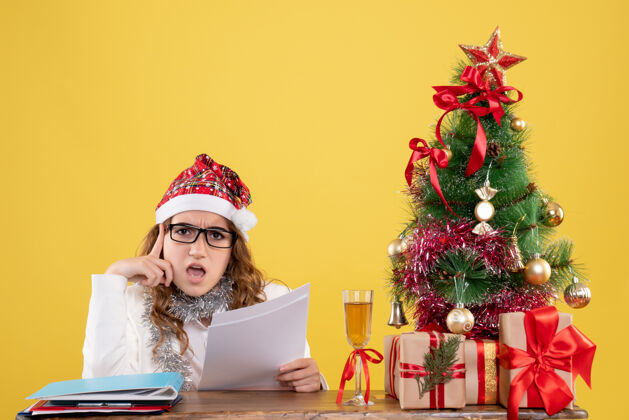 圣诞节前视图女医生坐在圣诞树上 拿着黄色背景上的文件圣诞树健康医生