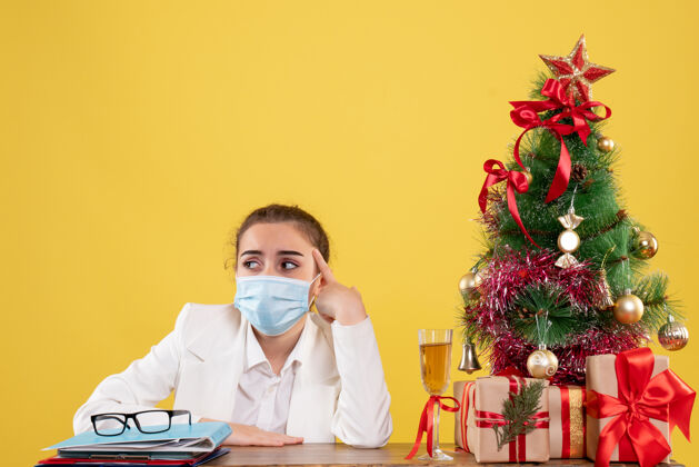 女性正面图女医生戴着防护面具坐在黄色背景上 带着圣诞树和礼品盒圣诞节专业健康
