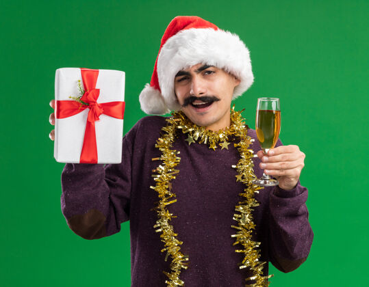香槟快乐的小胡子男人戴着圣诞老人的帽子 脖子上戴着金属丝 手里拿着一杯香槟和圣诞礼物 站在绿色的背景下 看着相机 开心地微笑着圣诞快乐圣诞脖子
