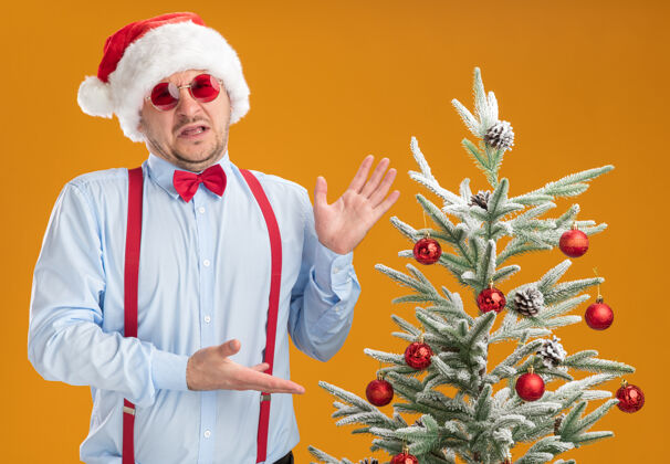 年轻人一个戴着圣诞帽 戴着红眼镜 系着吊带领结的年轻人站在圣诞树旁 用双臂把圣诞树抱在橘色的墙上 既恼怒又不高兴圣诞节帽子站着