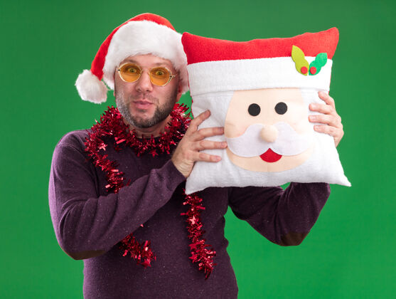 中年印象深刻的中年男子戴着圣诞帽 脖子上戴着金箔花环 戴着眼镜 把圣诞老人的枕头孤立在绿色的墙上枕头脖子印象深刻