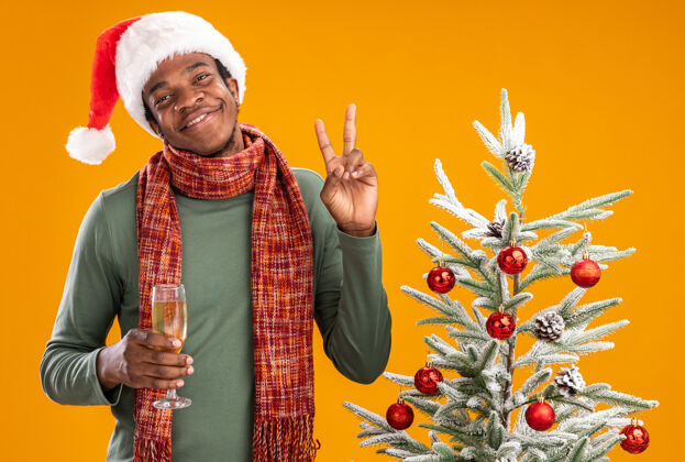围着戴着圣诞帽 脖子上围着围巾的非洲裔美国人 手里拿着一杯香槟 兴高采烈地微笑着 在橙色背景下 在圣诞树旁展示v形标志脖子圣诞树圣诞老人