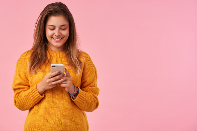 年轻快乐的年轻黑发可爱的女性 随意的发型 举着手机 带着淡淡的微笑积极的看着屏幕 孤立的粉红色手站立肖像