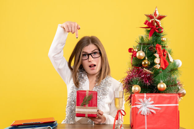 背景正面图：女医生坐在桌子前 拿着礼物 黄色背景上的圣诞树和礼盒肖像坐着漂亮