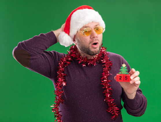 圣诞老人惊讶的中年男子戴着圣诞帽 脖子上戴着金箔花环 戴着眼镜 手里拿着圣诞树玩具玩具帽子花环