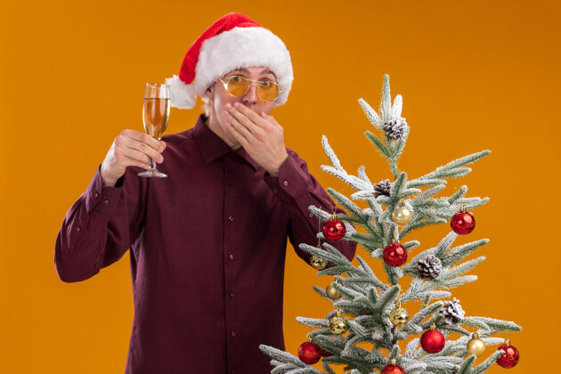圣诞老人一个戴着圣诞帽和眼镜的年轻金发男人站在橙色背景的装饰过的圣诞树旁手香槟站着