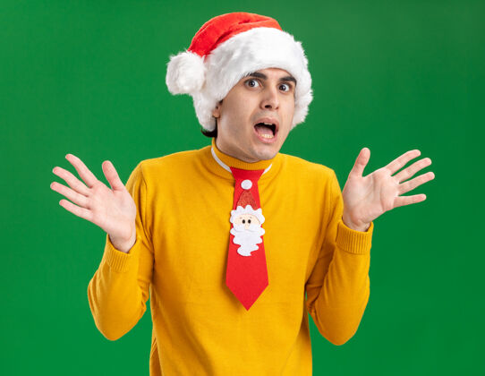 相机一个穿着黄色高领毛衣 戴着圣诞老人帽 打着滑稽领带的年轻人 站在绿色的背景上 一边高兴一边惊讶地看着镜头男人帽子圣诞老人