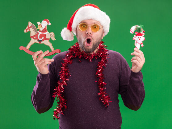 脖子惊讶的中年男子戴着圣诞帽 脖子上戴着金箔花环 戴着眼镜 手里拿着糖果藤饰品和圣诞老人穿着举行摇摆