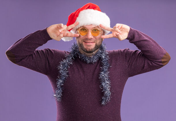 帽子高兴的中年男子戴着圣诞帽 脖子上戴着金属丝花环 戴着眼镜 看着相机上显示的v字符号穿着看Vsign
