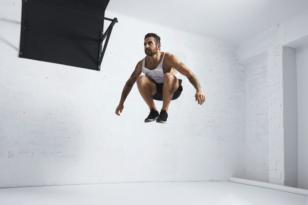 肌肉留着胡须和纹身的年轻男运动员在空中表演健美操跳高 隔离在健身中心的白色房间里成人器械年轻