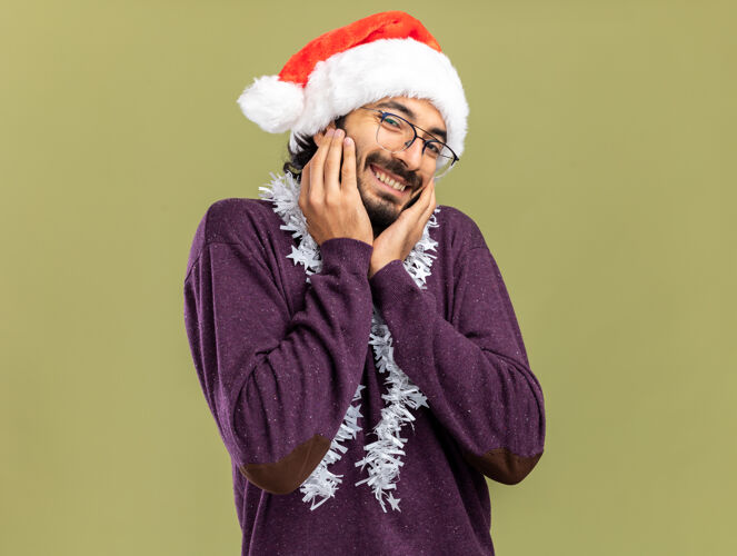脸颊带着微笑的年轻帅哥戴着圣诞帽 脖子上戴着花环 双手放在脸颊上 橄榄绿的背景是孤立的微笑帽子脖子