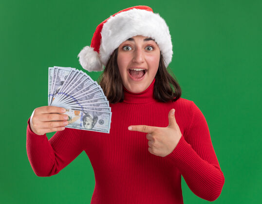 圣诞老人穿着红毛衣 戴着圣诞帽的年轻女孩拿着现金 用食指指着钱 站在绿色的墙上 又高兴又惊讶钱指着手指