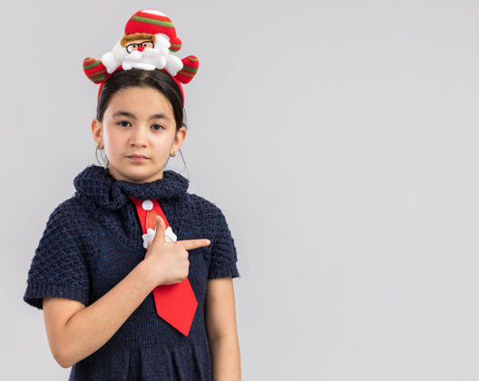 头小女孩穿着针织连衣裙 头上系着一条红色领带 戴着滑稽的圣诞戒指 表情严肃 手指指向一边女孩指向圣诞节