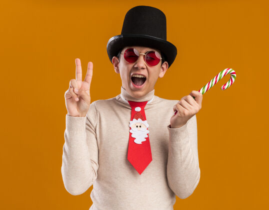 穿着兴奋的年轻人戴着帽子 戴着圣诞领带 戴着眼镜 手里拿着圣诞糖果 在黄色的墙上表现出一种与世隔绝的和平姿态帽子领带手势
