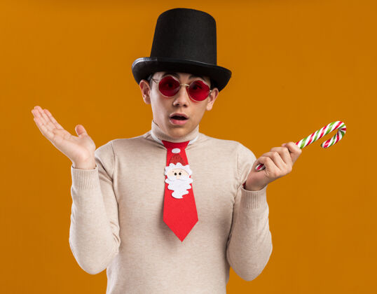 穿着困惑的年轻人戴着帽子 打着圣诞领带 戴着眼镜 手里拿着圣诞糖果 手被隔离在黄色的墙上帽子领带年轻