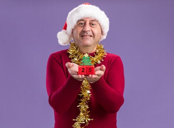 圣诞老人中年男子戴着圣诞老人的帽子 脖子上戴着金属丝 展示着玩具方块 日期是25岁 站在紫色的背景下 看着相机 开心地微笑着背景显示日期