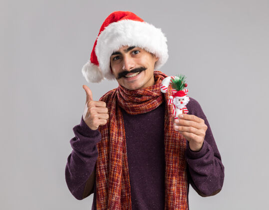 快乐快乐的小胡子男人戴着圣诞老人的帽子 脖子上围着暖和的围巾 手里拿着圣诞糖果手杖 微笑着站在白色背景上竖起大拇指看着摄像机站着糖果圣诞快乐