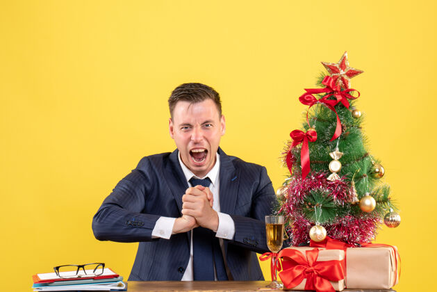 坐着前视图兴高采烈的男子坐在圣诞树旁的桌子上 呈现黄色礼物男人会议