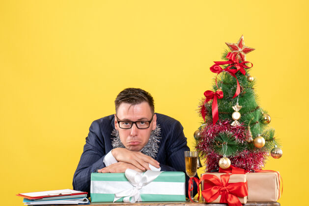 男人圣诞树旁坐在桌子旁的一个不快乐的男人的正视图 黄色的礼物执行官桌子电脑