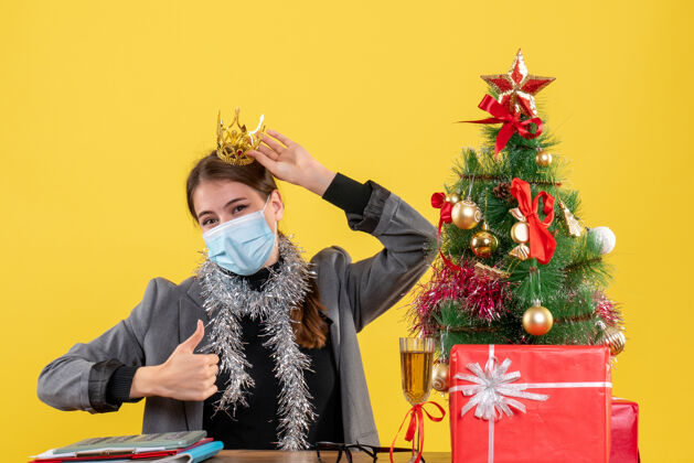 面具正面图戴着医学面具戴着皇冠的年轻女孩制作拇指向上标志圣诞树和礼物鸡尾酒拇指圣诞节礼物