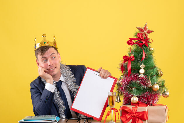 新郎戴着皇冠的帅哥坐在圣诞树旁的桌子前 黄色的礼物笔记本电脑黄色帅哥