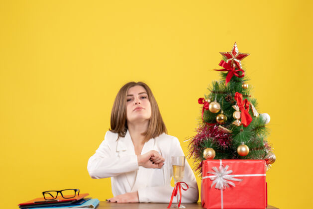 女性正面图：女医生坐在黄色背景的桌子前 放着圣诞树和礼品盒前面微笑插花