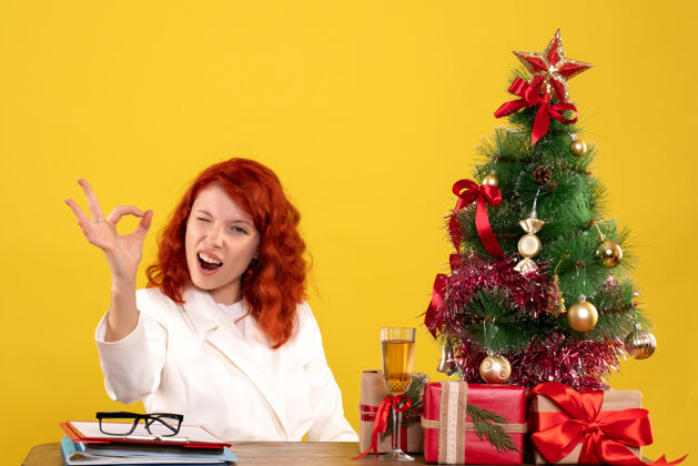 桌子正面图：女医生坐在桌子后面 拿着圣诞礼物 黄色背景上的树女医生圣诞节情感