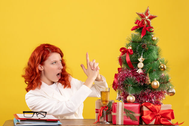 肖像前视图：女医生坐在桌子后面 黄色背景上有圣诞礼物人成人礼物