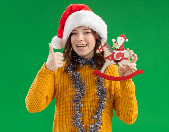 女孩快乐的年轻斯拉夫女孩戴着圣诞帽 脖子上戴着花环 把圣诞老人抱在摇马装饰上 竖起大拇指周围斯拉夫举行