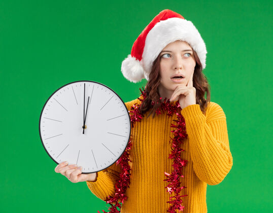 时钟戴着圣诞帽 脖子上戴着花环的年轻斯拉夫女孩惊讶地把手指放在脸上 拿着时钟看着一边绿色举行复制