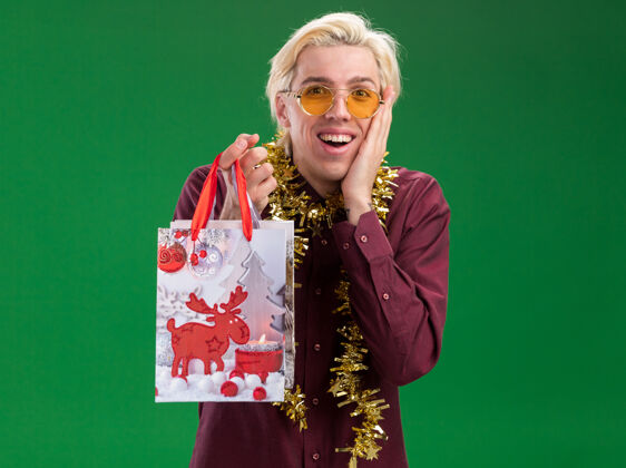 男人兴奋的年轻金发男子戴着眼镜 脖子上戴着金属丝花环 手里拿着圣诞礼品袋 手放在脸上 隔离在绿色的墙上 留着复印空间周围花环保持
