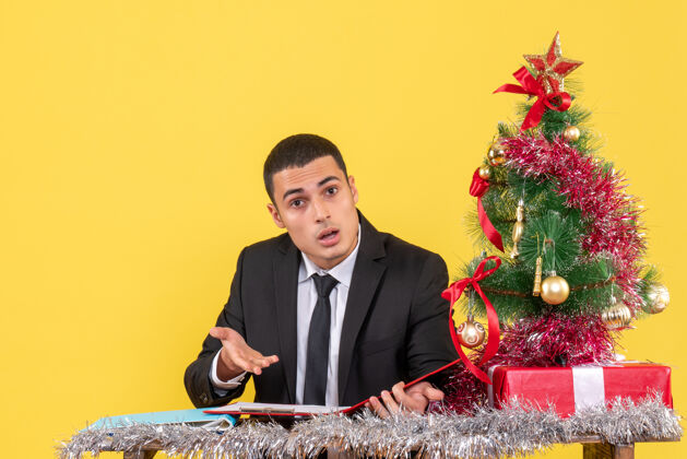 公司前视图困惑的西服男子坐在桌子旁拿着文件圣诞树和礼物坐着男人桌子