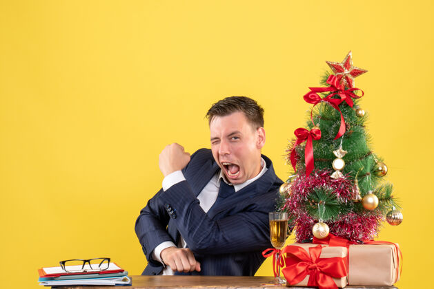赢一个英俊的男人坐在圣诞树旁的桌子前展示胜利的姿态 黄色的礼物圣诞节演讲者西装