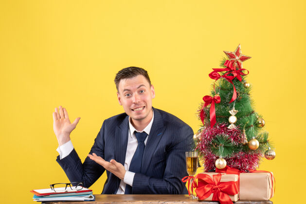 圣诞节正面是一个快乐的年轻人指着背景 坐在圣诞树旁的桌子上 黄色的礼物指点树执行
