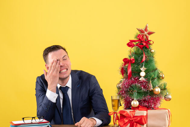 树哭泣的男人用手捂着眼睛坐在圣诞树旁的桌子前 黄色的礼物男人行政人员桌子
