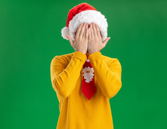年轻戴着黄色高领毛衣和圣诞帽的年轻人 戴着滑稽的领带 双手站在绿色背景上遮住眼睛手站着高领毛衣