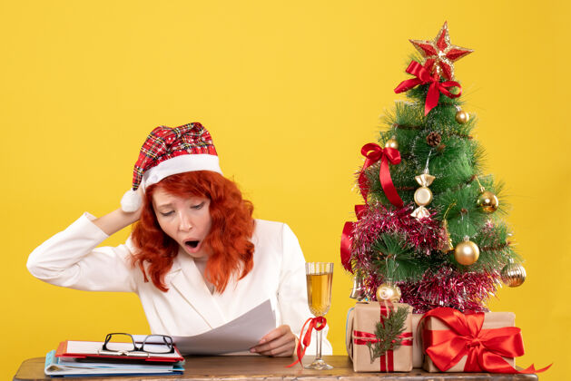 礼物前视图：女医生坐在桌子后面 黄色背景上有礼物 圣诞树和礼品盒医生背景帽子