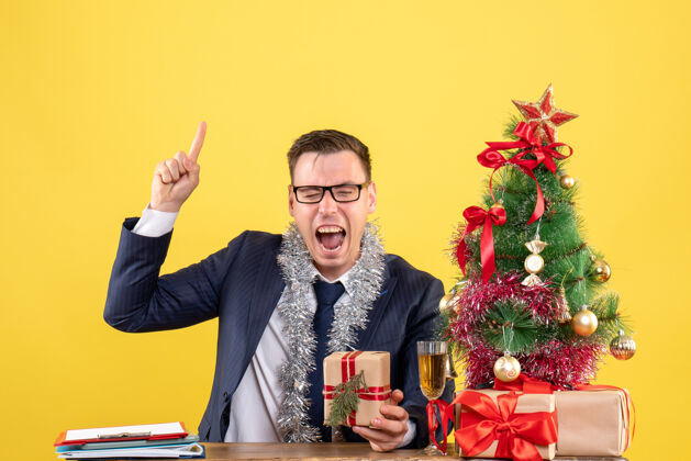 桌子前视图兴高采烈的男子手指庞起坐在桌旁圣诞树和黄色的礼物手指坐着行政人员