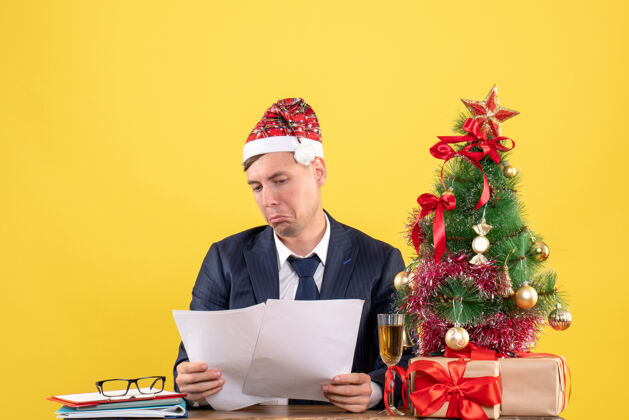 看不快乐的男人坐在圣诞树旁的桌子上看报纸 黄色的礼物桌子商人人