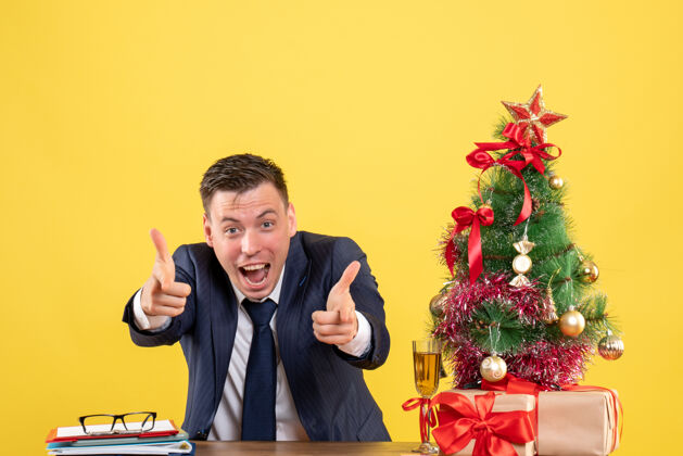 指着前视图兴高采烈的男子手指相机坐在圣诞树附近的桌子上 黄色的礼物行政人员办公室树