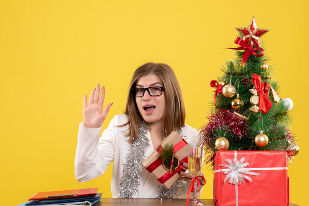 女性正面图：女医生坐在黄色背景的桌子前 放着圣诞树和礼品盒漂亮背景坐着