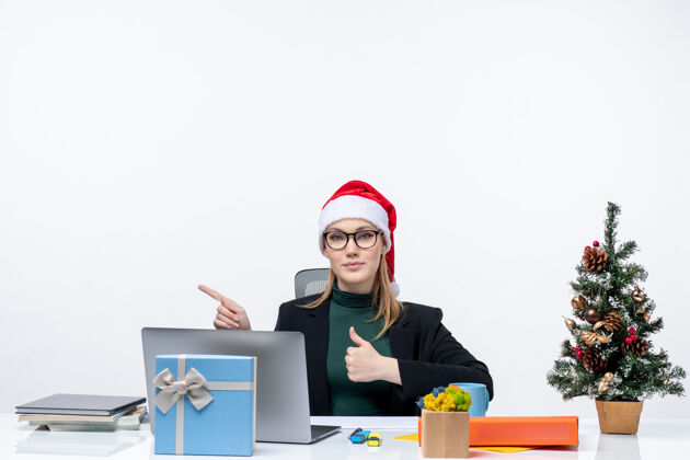 电脑戴着圣诞老人帽的女商人坐在一张桌子旁 桌上放着圣诞树和礼物圣诞老人好的右边