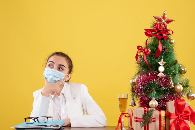 外套正面图女医生戴着防护面具坐在黄色背景上思考着圣诞树和礼品盒前面医疗人