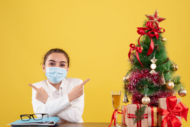 圣诞节正面图：女医生戴着防护面具坐在黄色背景上微笑 戴着圣诞树和礼盒女性女医生人