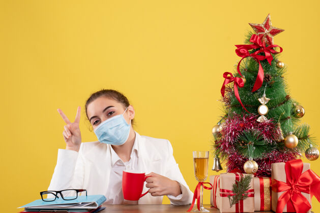 坐着正面图女医生戴着防护面具坐在黄色背景上 戴着圣诞树和礼盒人病毒防护