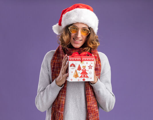 年轻快乐而积极的年轻人戴着圣诞老人帽 戴着黄色眼镜 脖子上围着暖和的围巾 手里拿着圣诞礼物 面带微笑地看着相机站在紫色的背景下微笑礼物背景