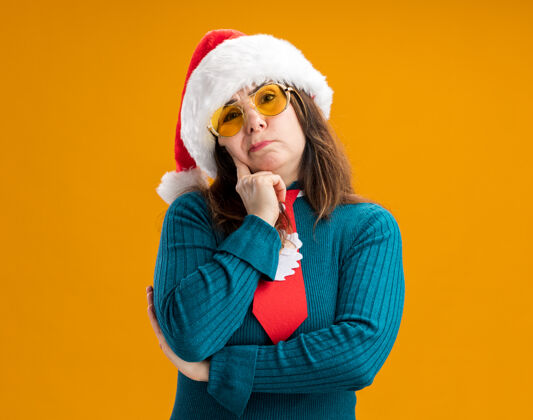 成人戴着太阳眼镜 戴着圣诞帽 系着圣诞领带的体贴的成年白人妇女把手指放在脸上 看着隔离在橙色背景上的相机 还有复印空间体贴领带圣诞老人
